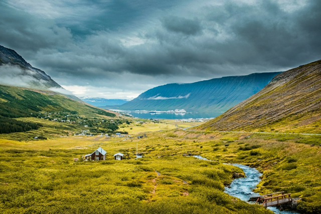 Scenic landscape of Ísafjörður, Iceland, showing a small village with scattered houses in a green valley, winding river in foreground, surrounded by steep mountains under dramatic dark cloudy sky, with fjord visible in the distance.
