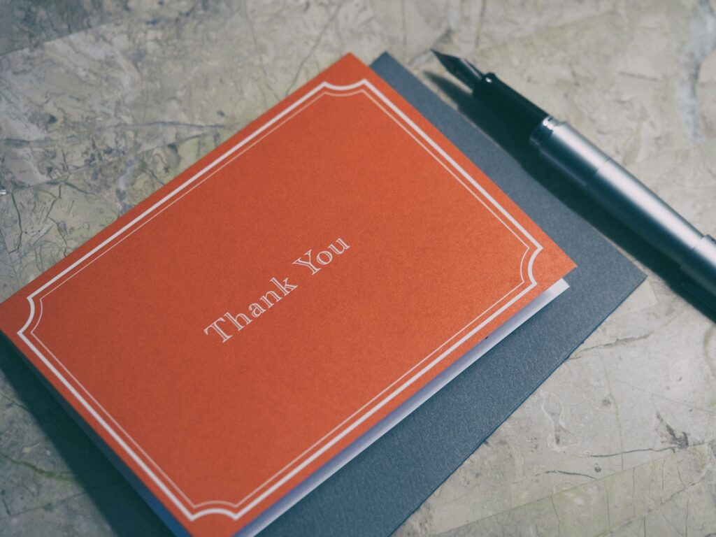 Close-up of a red “Thank You” card with white decorative border text, placed on a marble surface next to a black fountain pen, conveying gratitude, stationery, and elegant correspondence.