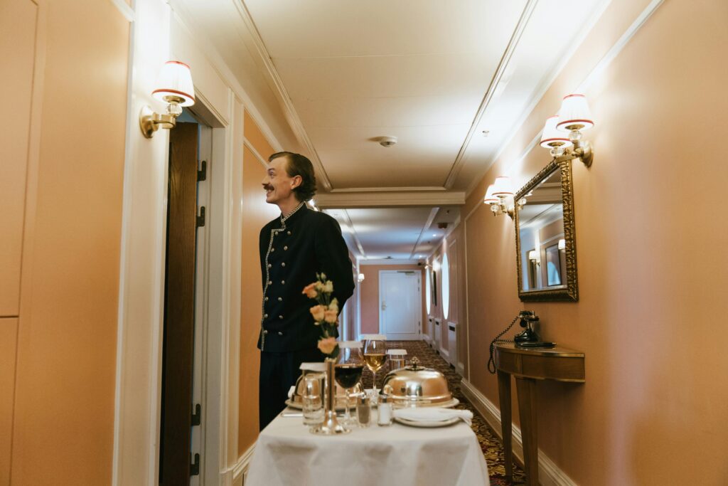 Cruise ship staff member in uniform delivering room service in a narrow cabin hallway, pushing a cart with covered dishes, wine glasses, and flowers, surrounded by warm lighting, wall sconces, mirror, and classic decor.